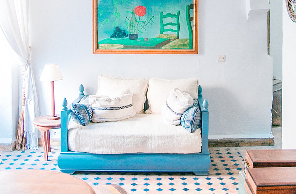 Bedroom with a  blue bed frame and white sheets and pillows with blue painting hanging over it on the wall. Small wooden night stand by the bed with a white lamp on it.