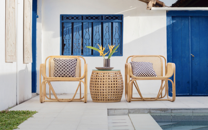 Two brown wooden chairs witha small stand inbetween with a plant on top infront of a house with a blue door and window